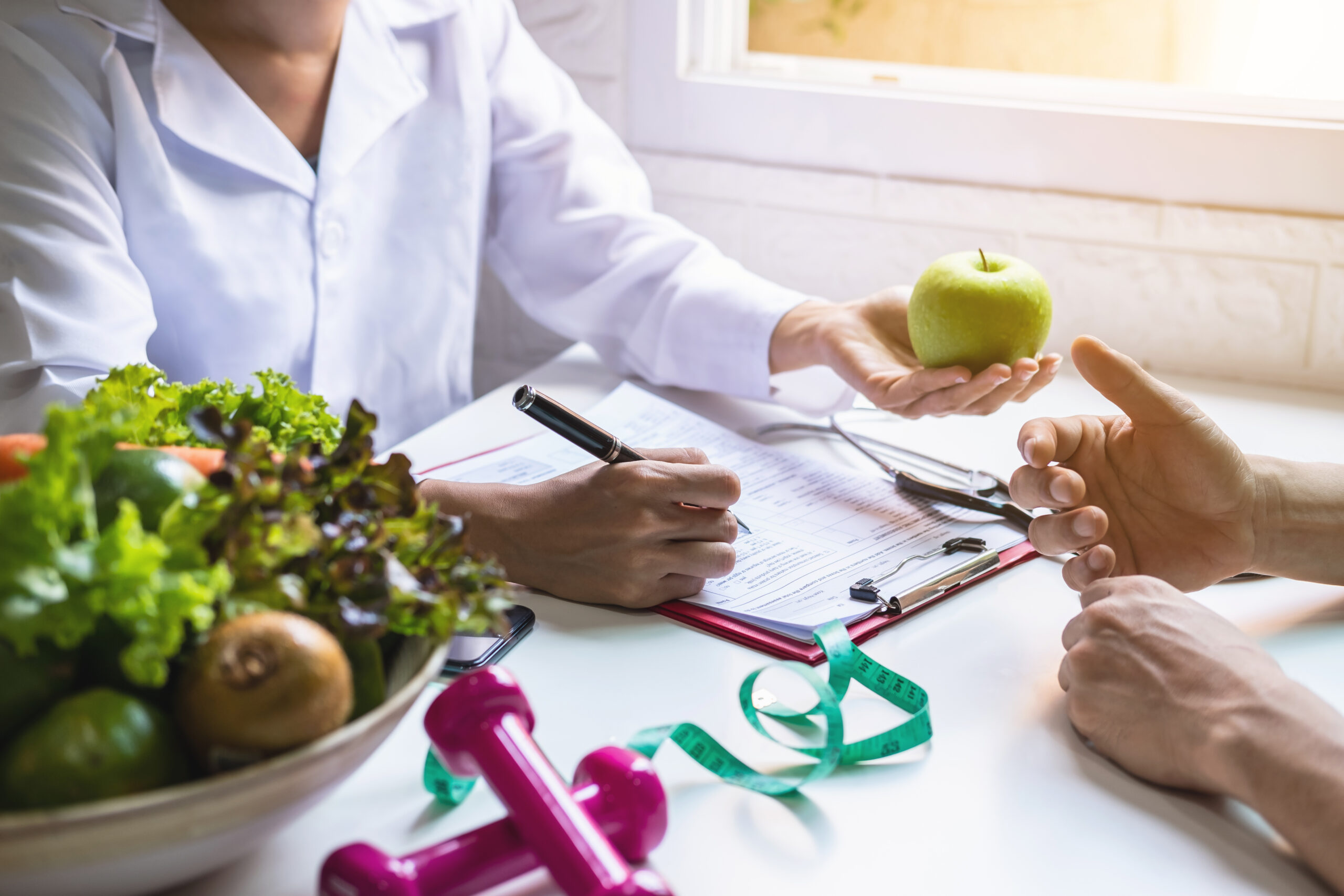 Ernährungsberater im Gespräch mit einem Klienten, der einen Apfel hält, mit Fokus auf gesunde Lebensmittel und Trainingsutensilien im Vordergrund Ernährungsberater im Gespräch mit einem Klienten, der einen Apfel hält, mit Fokus auf gesunde Lebensmittel und Trainingsutensilien im Vordergrund